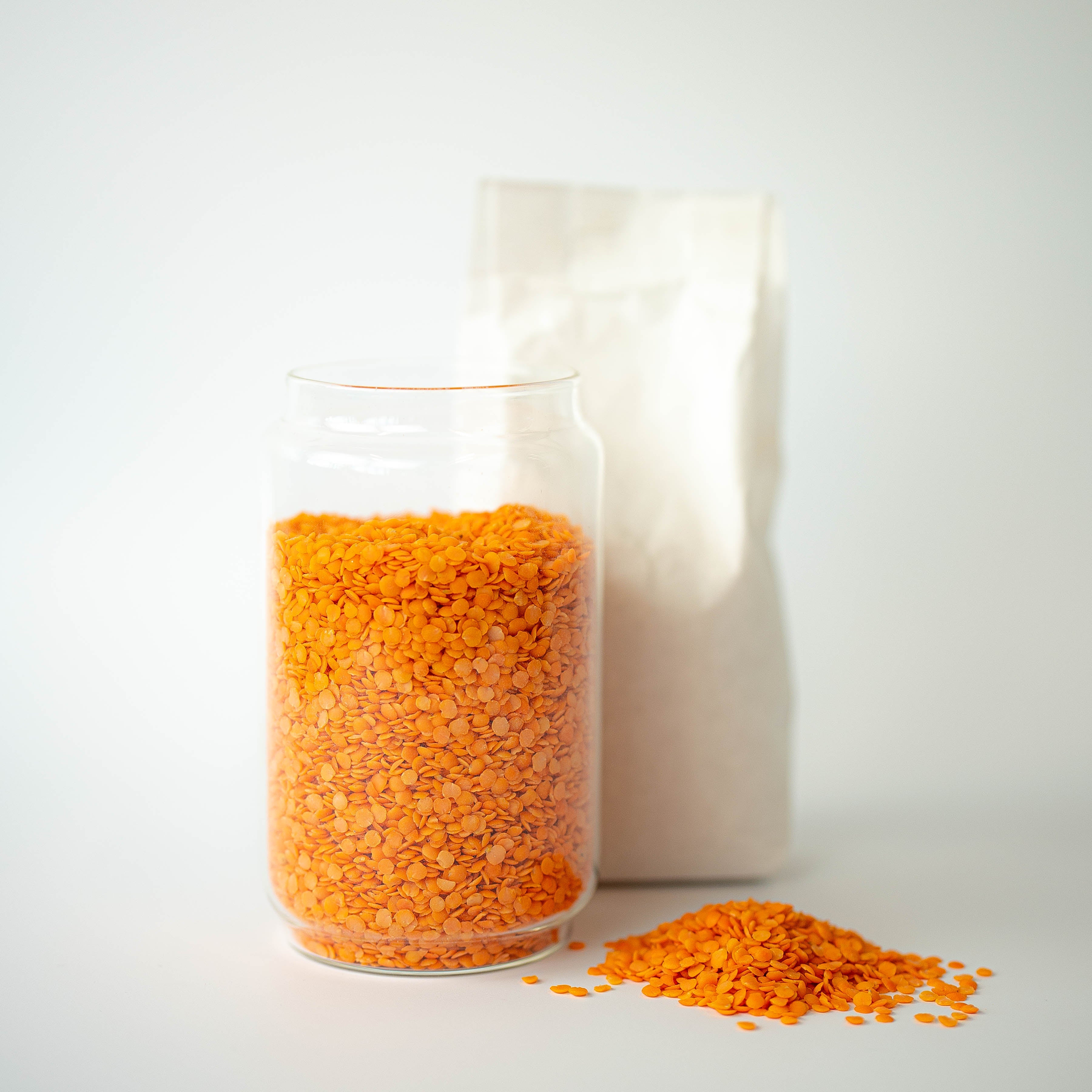 A clear glass jar filled with organic red split lentils, with a recyclable paper bag in the background and a small pile of lentils on the foreground.