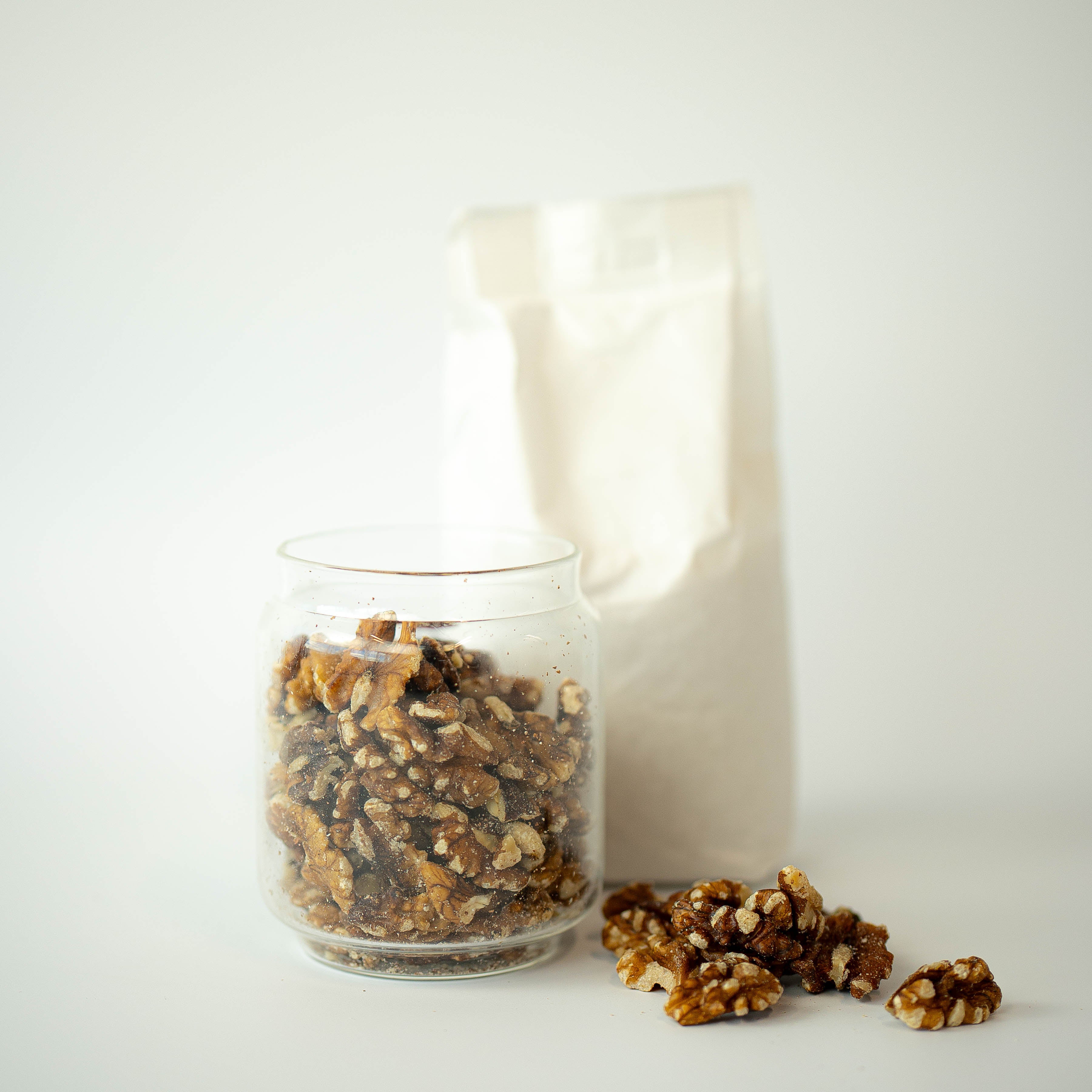 A glass jar filled with walnut halves next to a recyclable paper bag, with some walnuts scattered in front.