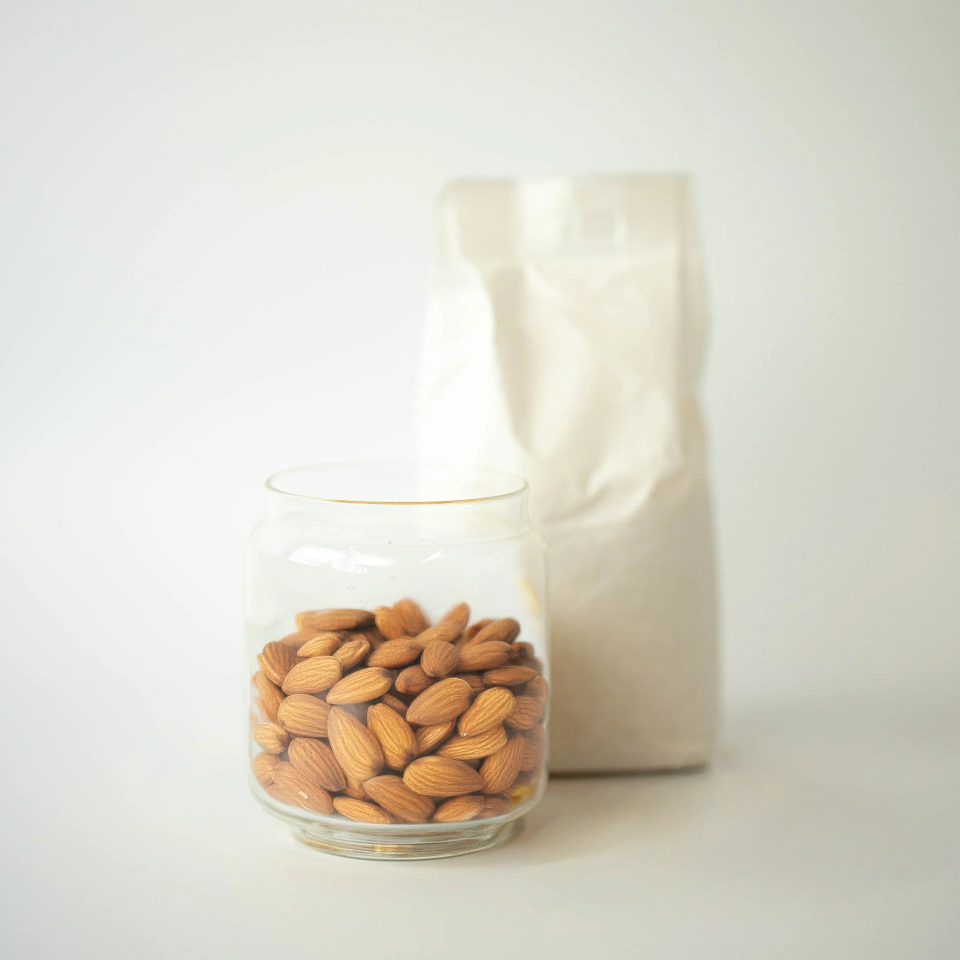 A glass jar filled with whole almonds, with a recyclable paper bag of almonds in the background.