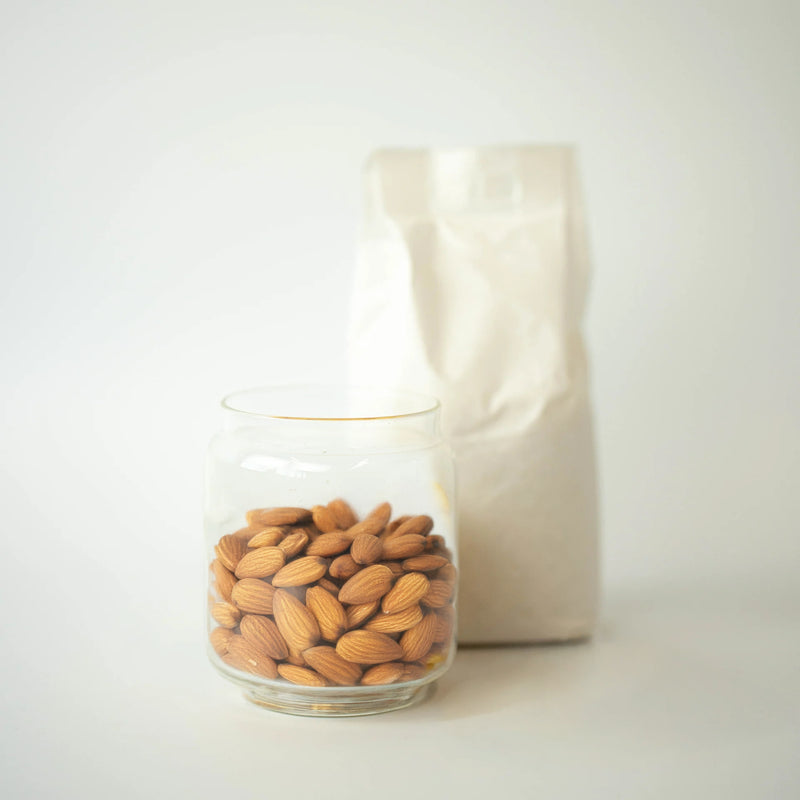 A glass jar filled with whole almonds, with a recyclable paper bag of almonds in the background.