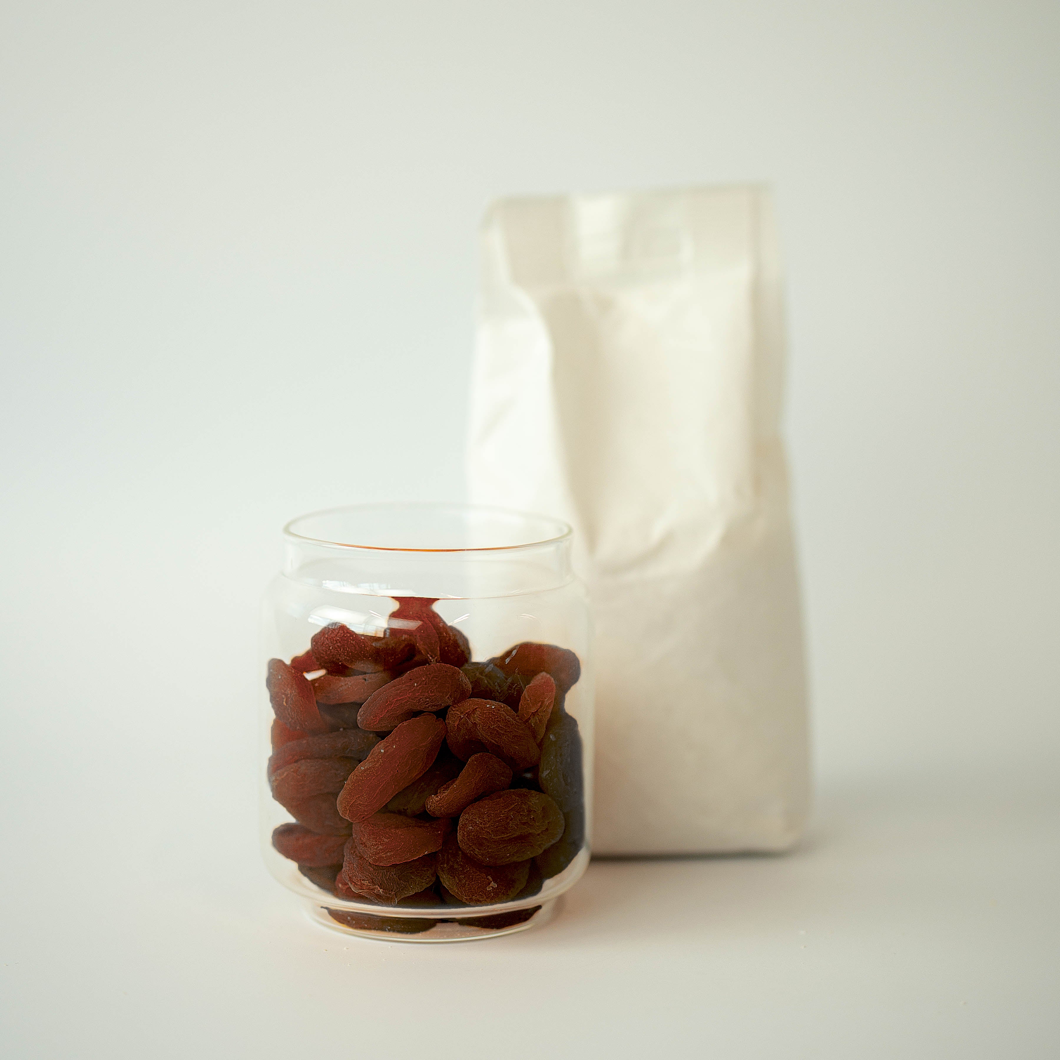Glass jar filled with dried apricots next to a white bag on a light background