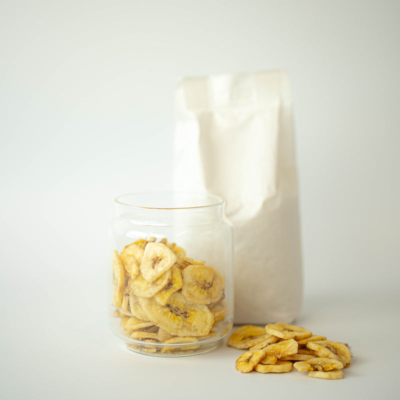 A glass jar filled with organic banana chips in front of a recyclable paper bag, with some chips scattered on the surface.