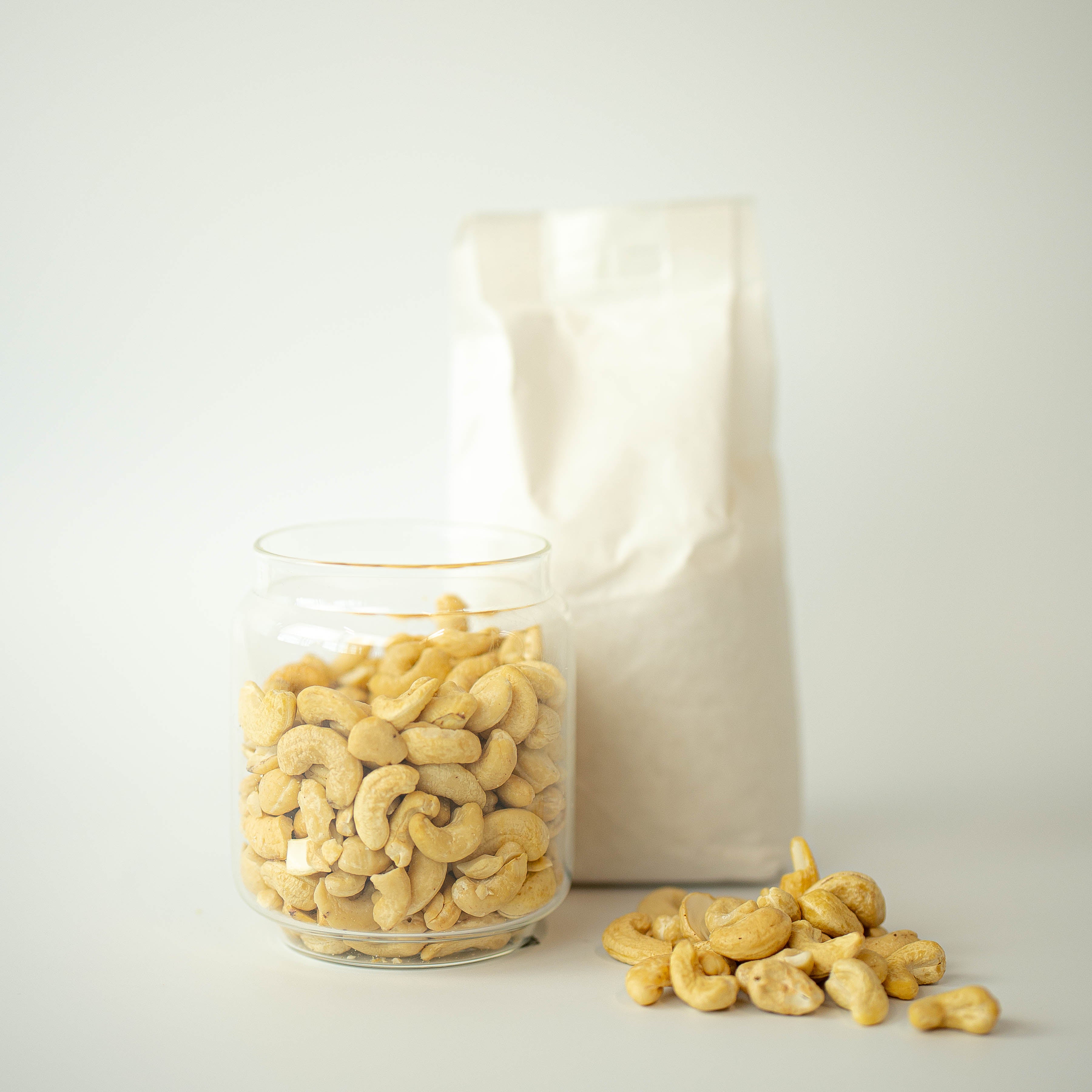 A glass jar filled with cashew nuts is placed in front of a recyclable paper bag, with some cashew nuts scattered in the foreground.