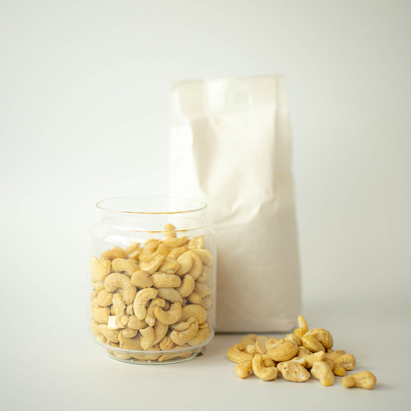 A glass jar filled with cashew nuts is placed in front of a recyclable paper bag, with some cashew nuts scattered in the foreground.