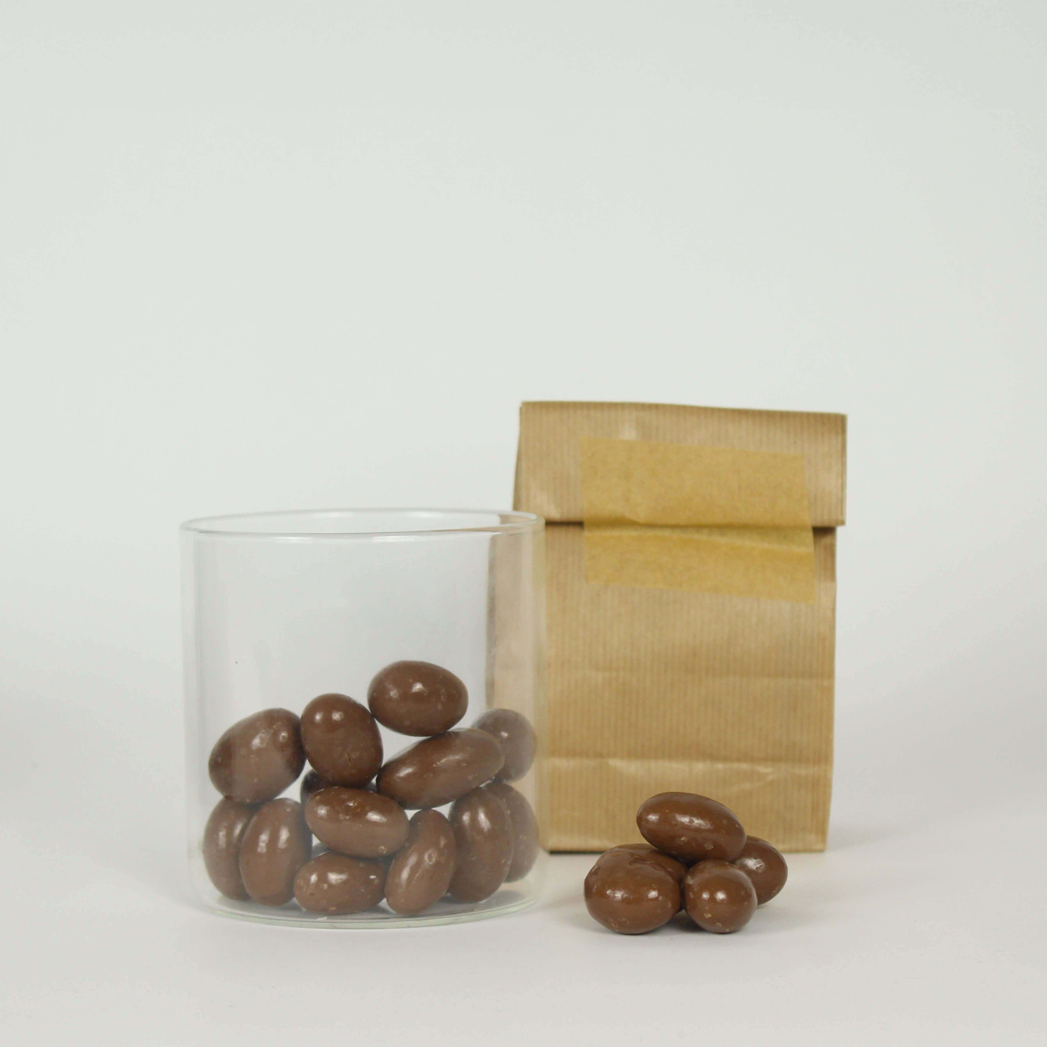 Glass jar and brown paper bag with chocolate-covered Brazil nuts on a white background