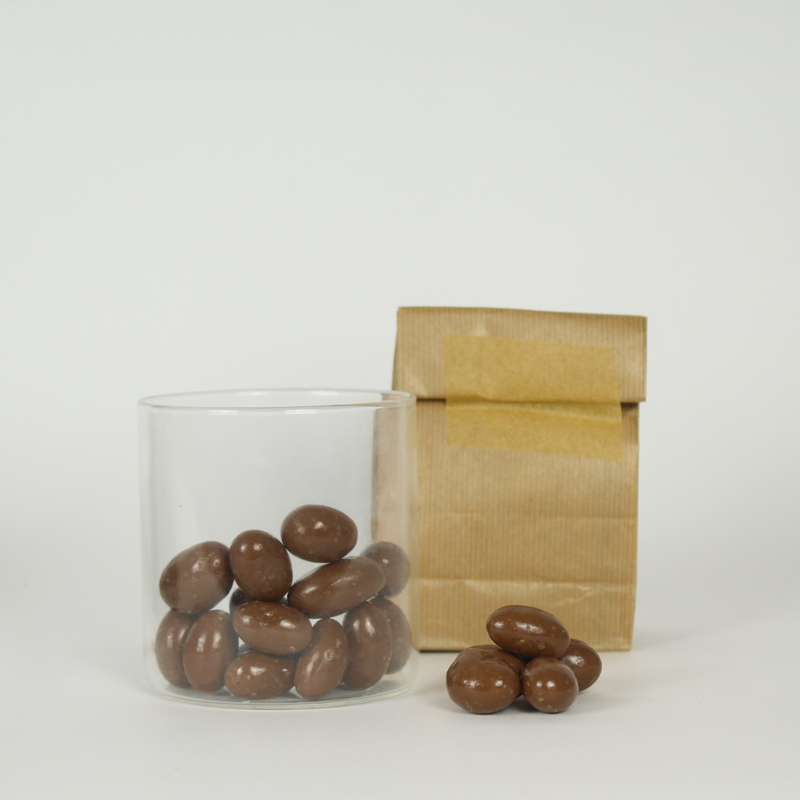 Glass jar and brown paper bag with chocolate-covered Brazil nuts on a white background