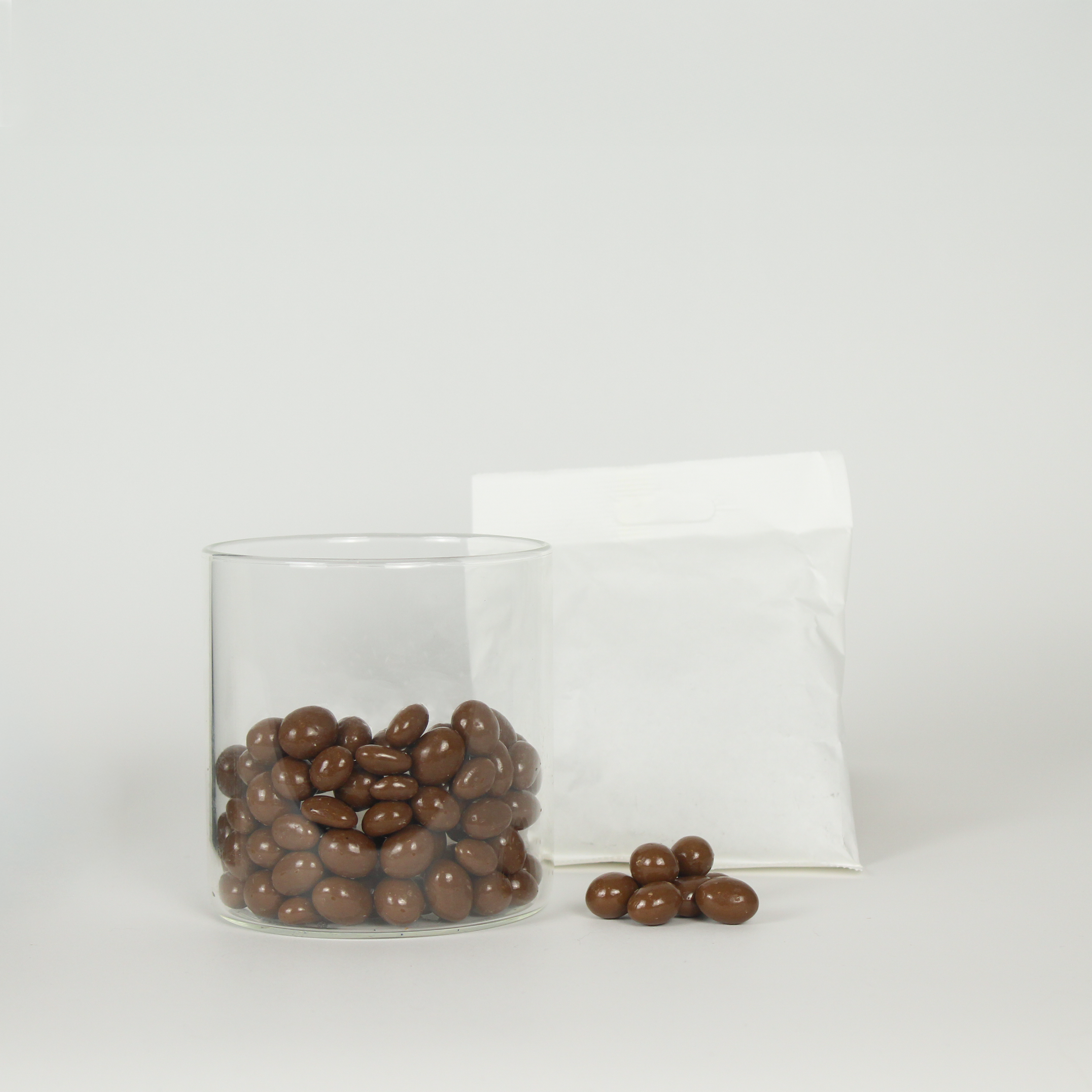 Glass container filled with chocolate-covered raisins next to a white bag on a light gray background