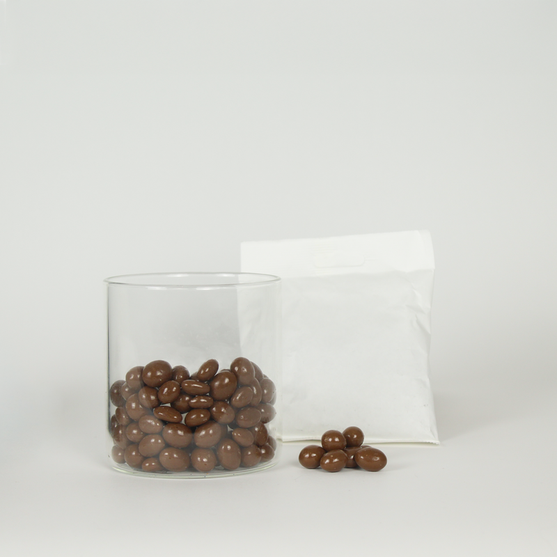 Glass container filled with chocolate-covered raisins next to a white bag on a light gray background