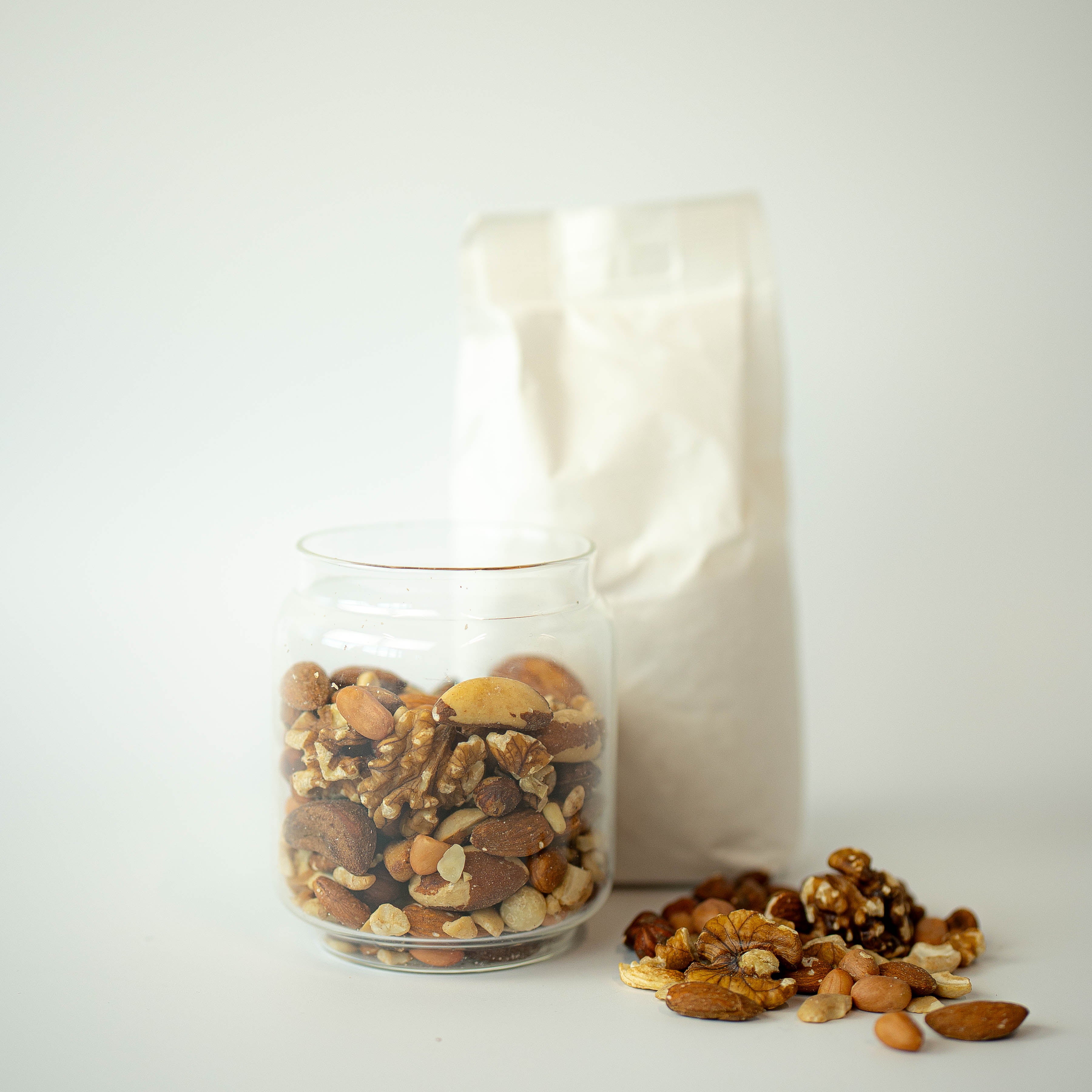 A glass jar filled with mixed nuts, with a recyclable paper bag in the background, and a scattering of nuts on the surface.