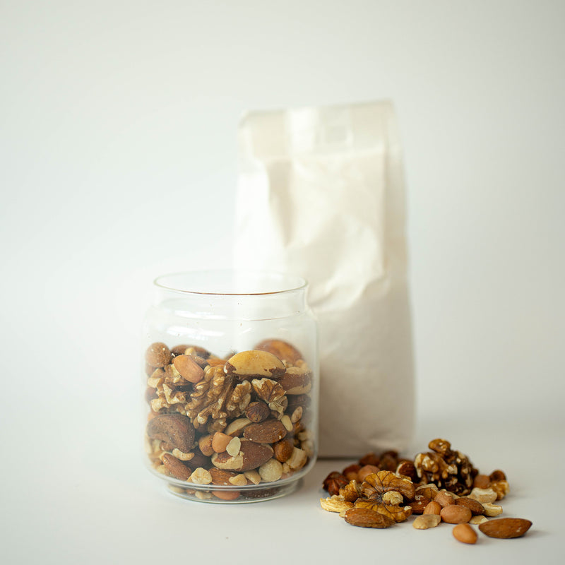 A glass jar filled with mixed nuts, with a recyclable paper bag in the background, and a scattering of nuts on the surface.