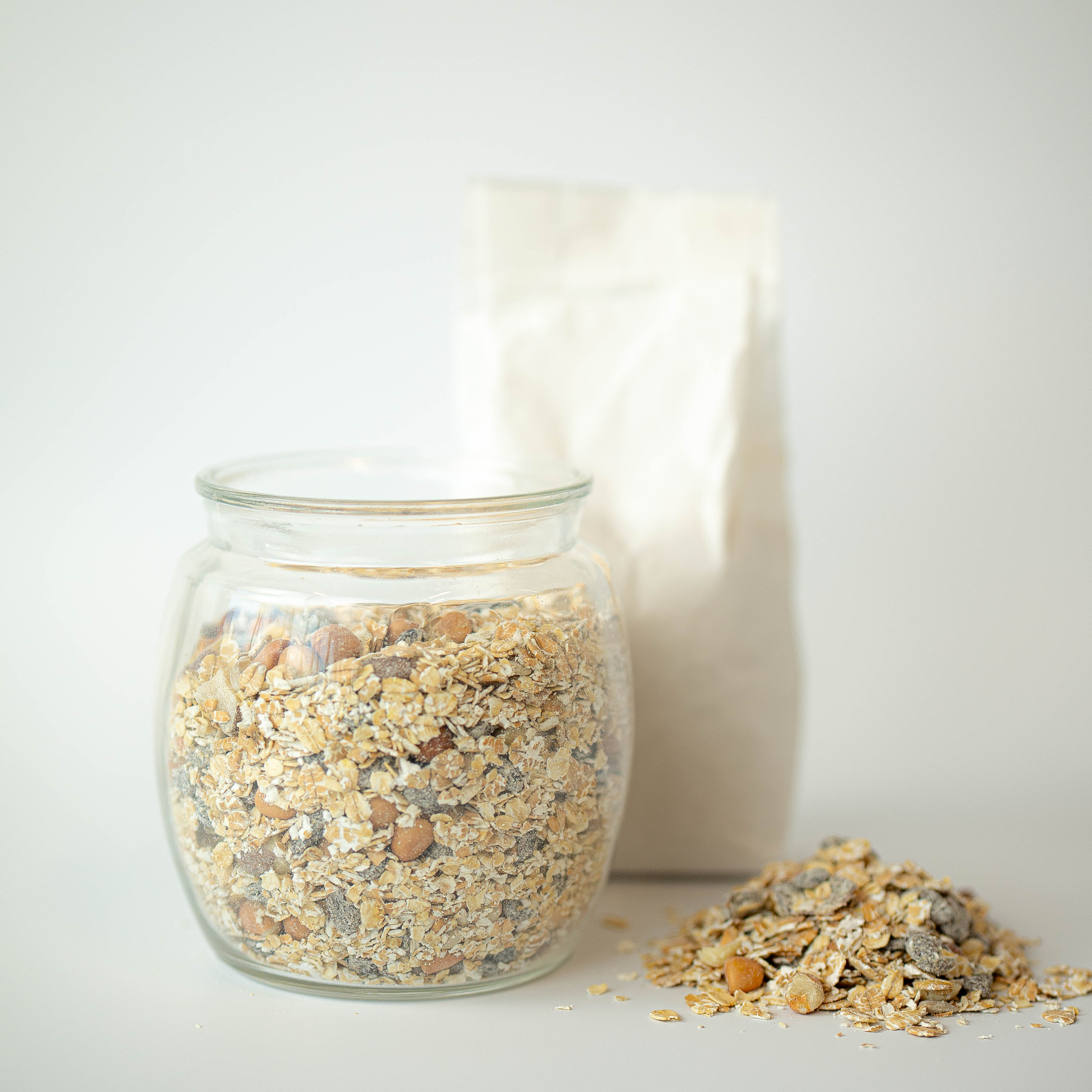 A glass jar filled with muesli cereal, with a recyclable paper bag in the background and a pile of muesli cereal in the foreground.