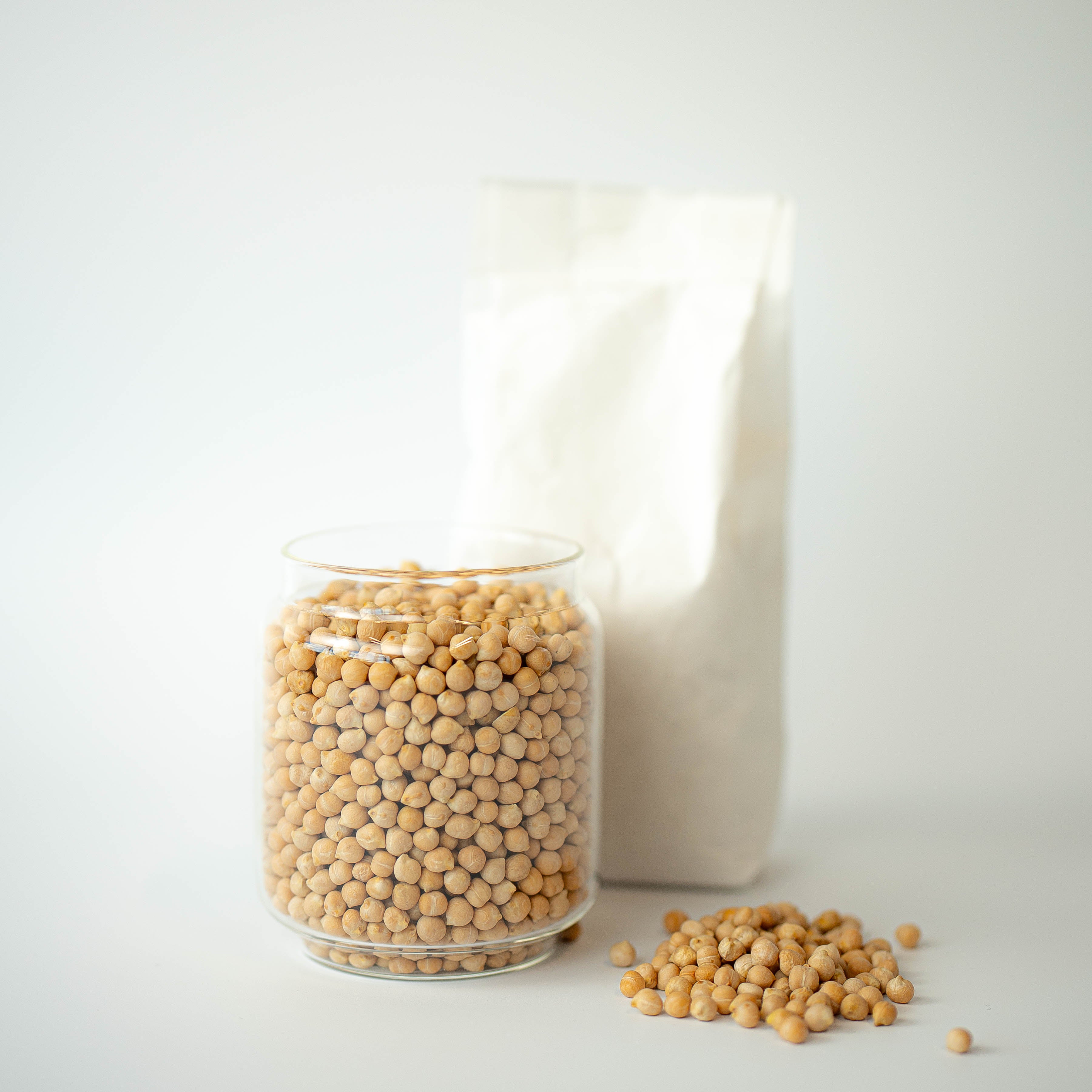 A glass jar filled with organic dried chickpeas is placed in front of a recyclable paper bag, with some chickpeas scattered on the surface.