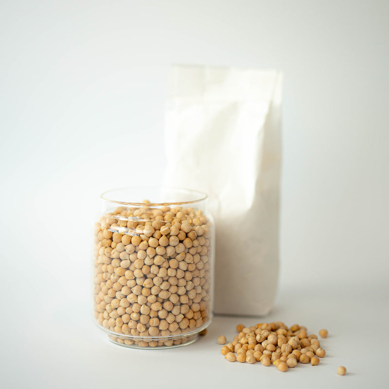 A glass jar filled with organic dried chickpeas is placed in front of a recyclable paper bag, with some chickpeas scattered on the surface.