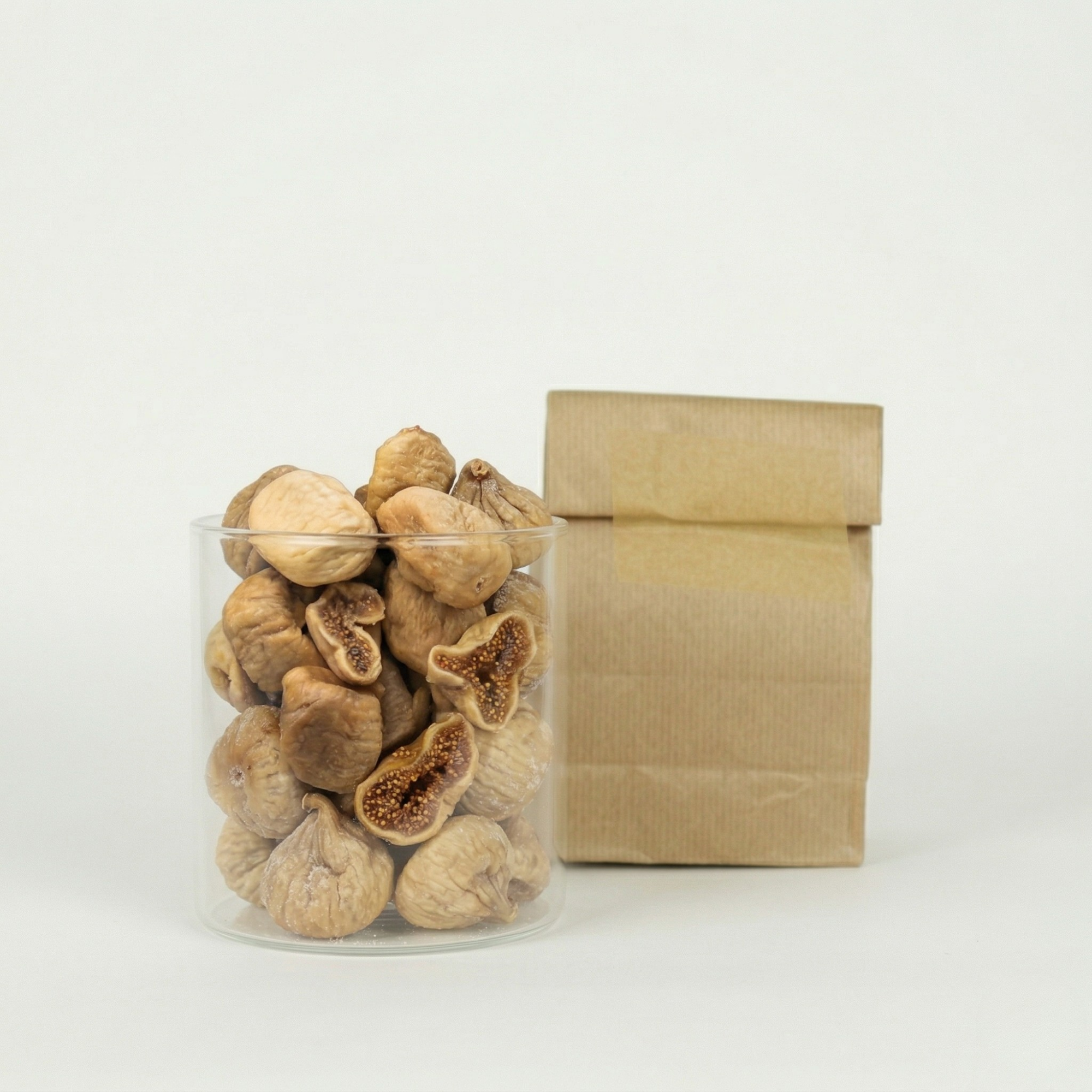 Clear glass jar filled with dried figs next to a brown paper bag on a white background