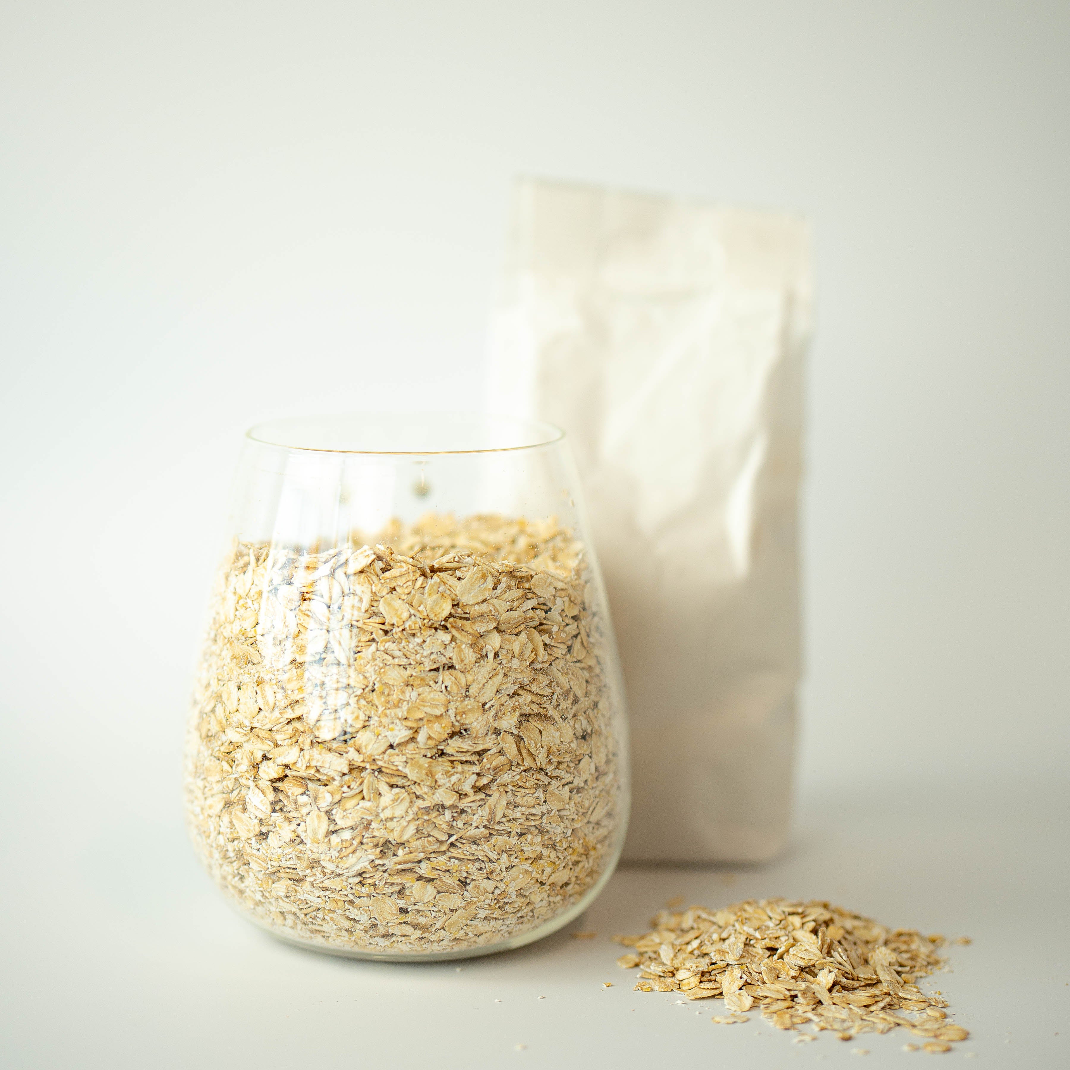 A glass jar filled with jumbo oatflakes, with a recyclable paper bag in the background and a small pile of oatflakes in the foreground.