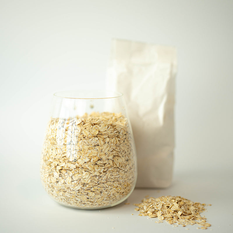 A glass jar filled with jumbo oatflakes, with a recyclable paper bag in the background and a small pile of oatflakes in the foreground.