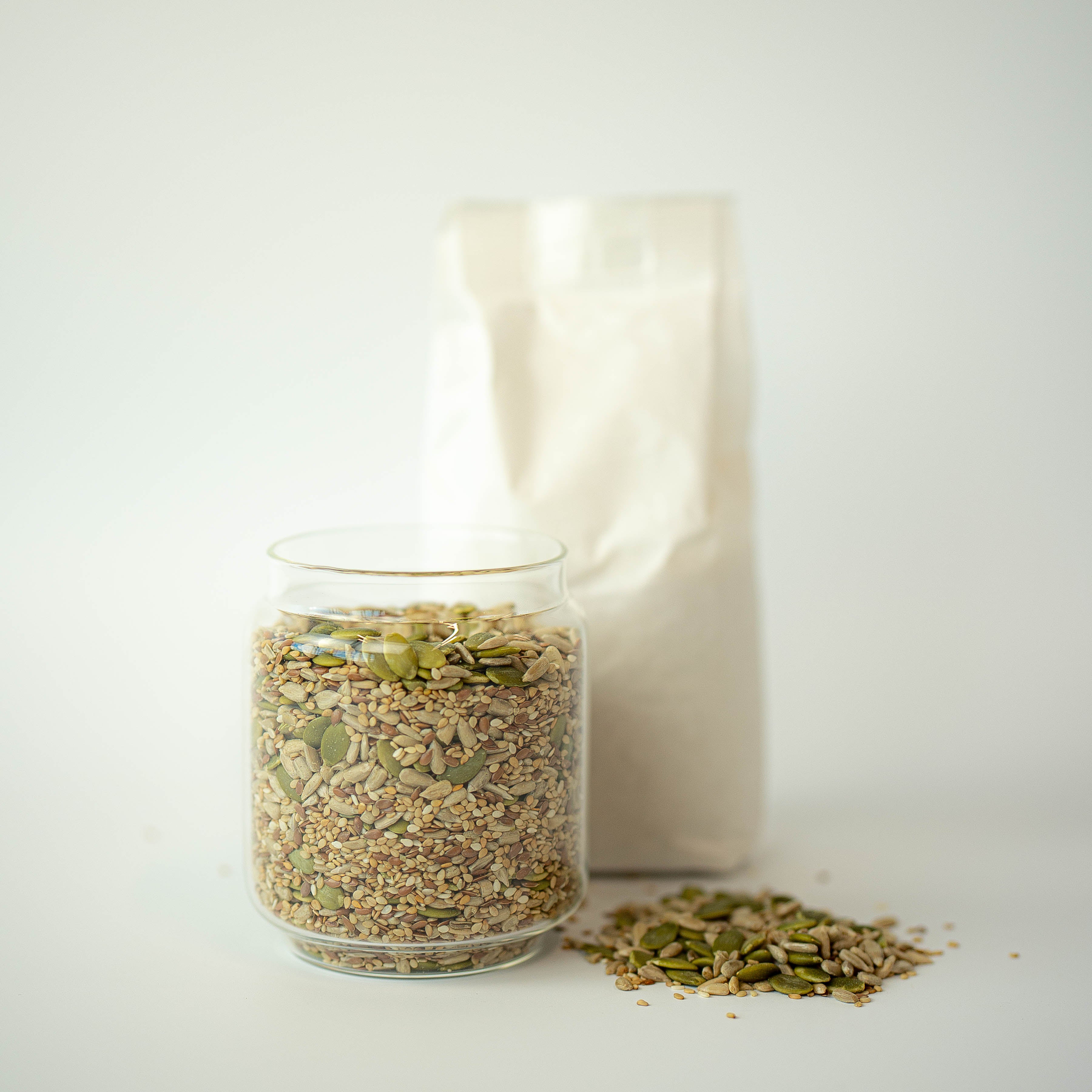 A glass jar filled with omega seed mix, in front of a recyclable paper bag, with a small amount of seeds scattered on the surface beside it.