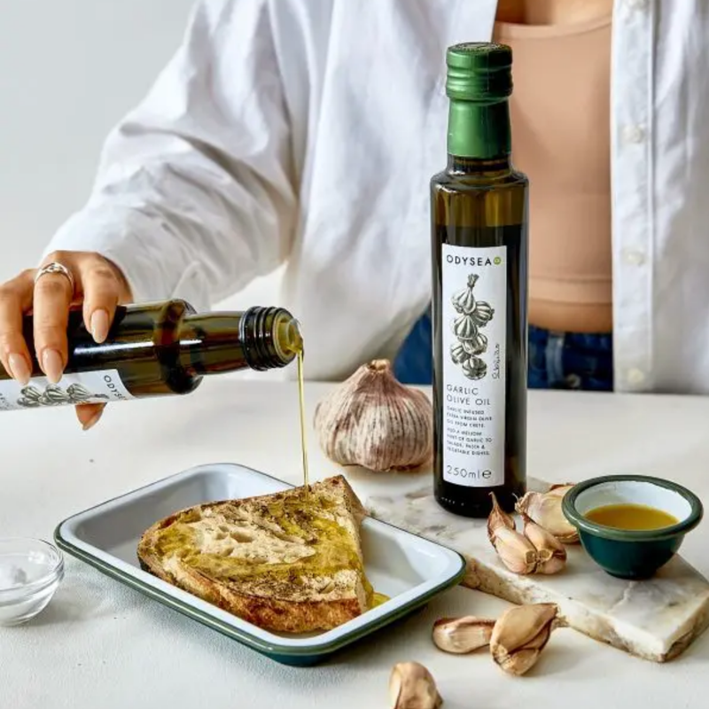 Person pouring odysea garlic olive oil from a bottle onto bread with garlic and olive oil on a table.