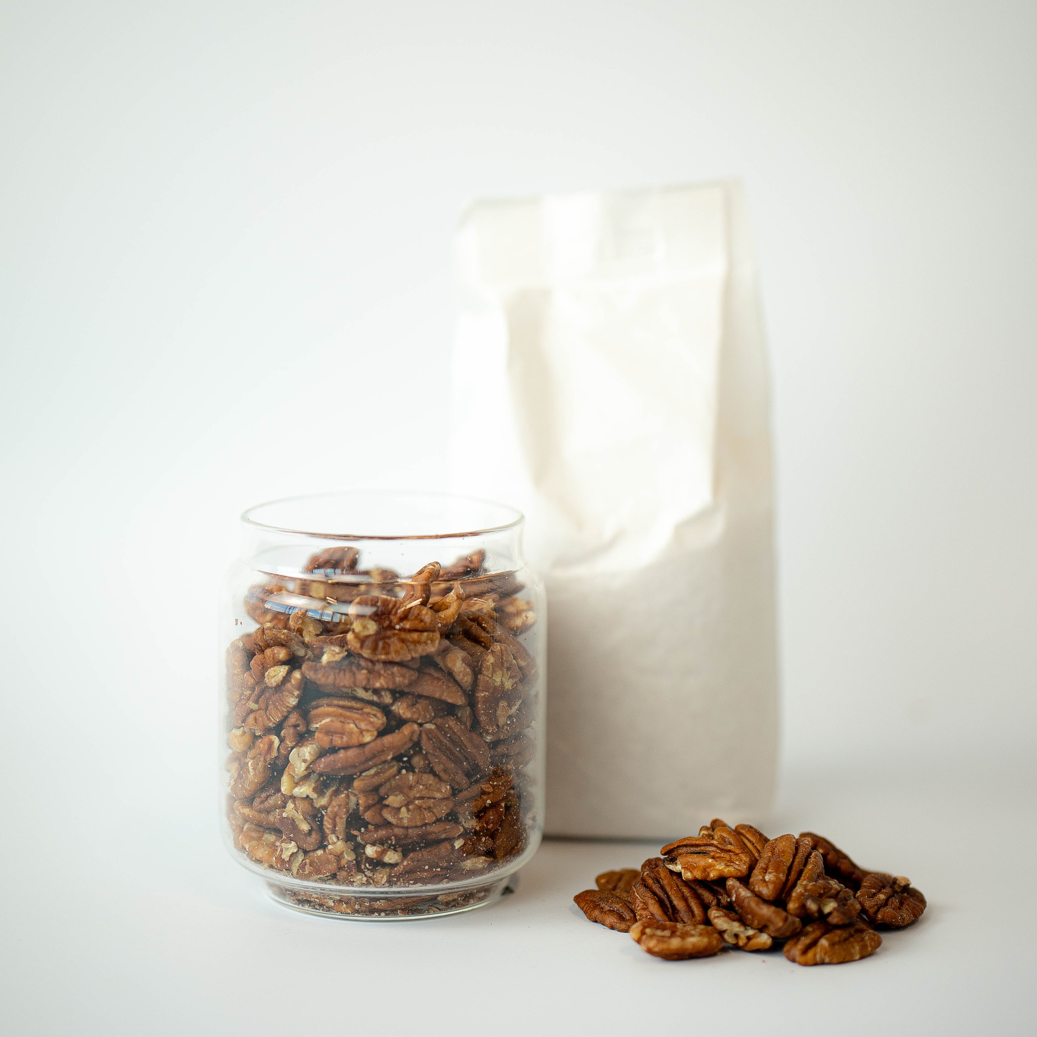 A glass jar filled with pecan halves next to a recyclable paper bag, with some pecan halves scattered in front.