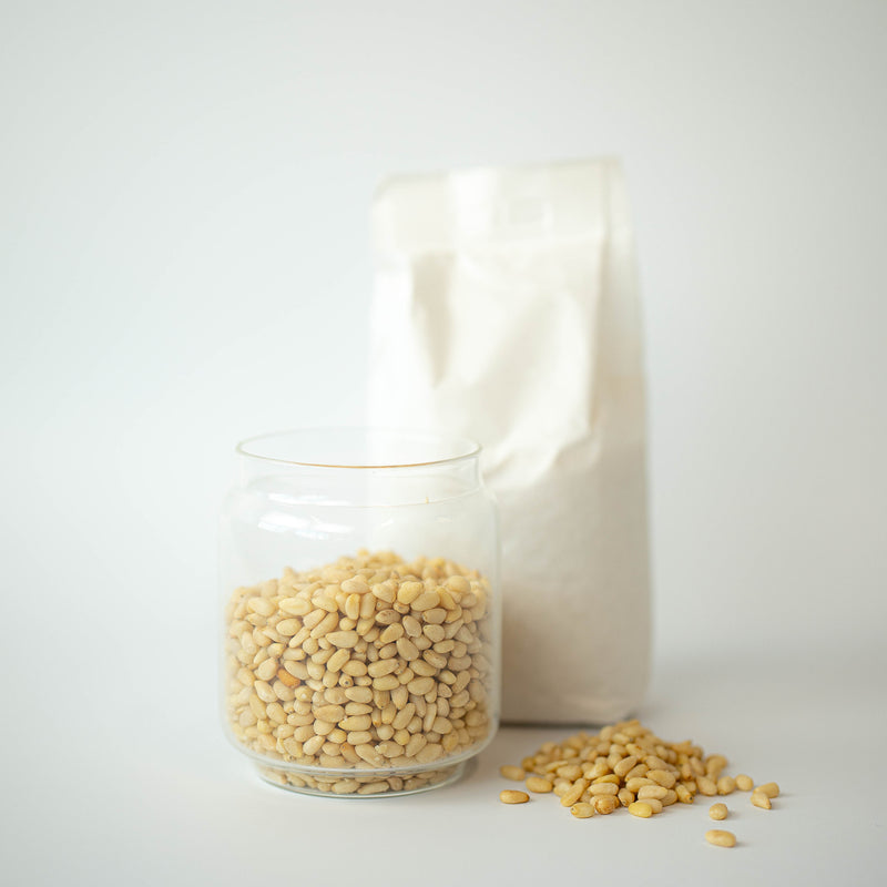 A glass jar filled with pine nuts in front of a recyclable paper bag, with some pine nuts scattered on the surface.
