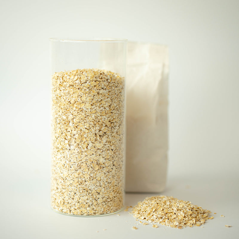 A clear jar filled with porridge oats in the foreground, with a recyclable paper bag partially visible in the background. A small pile of oats is also visible in front of the jar.