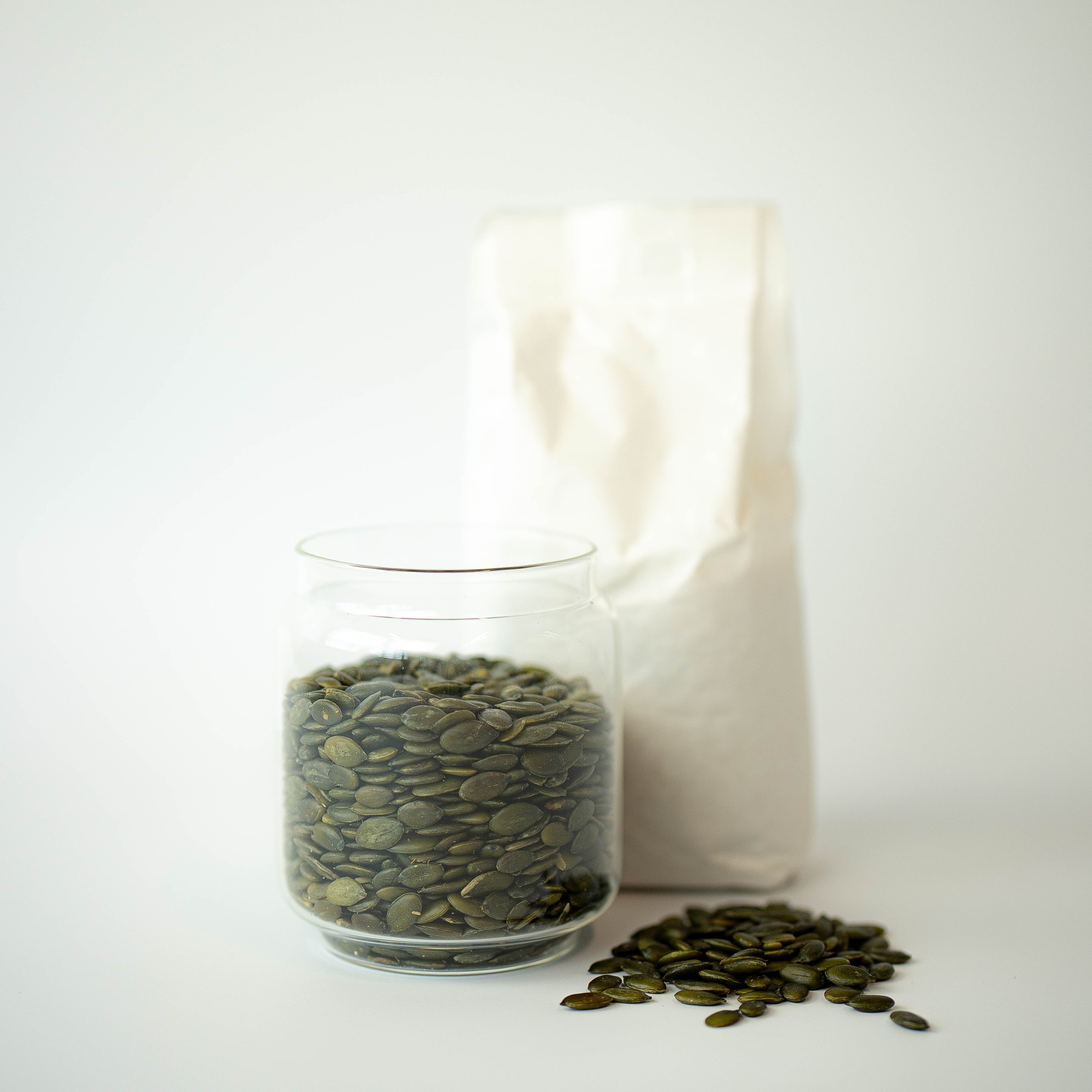 A glass jar filled with organic pumpkin seeds in front of a recyclable paper bag, with a few seeds scattered in the foreground.