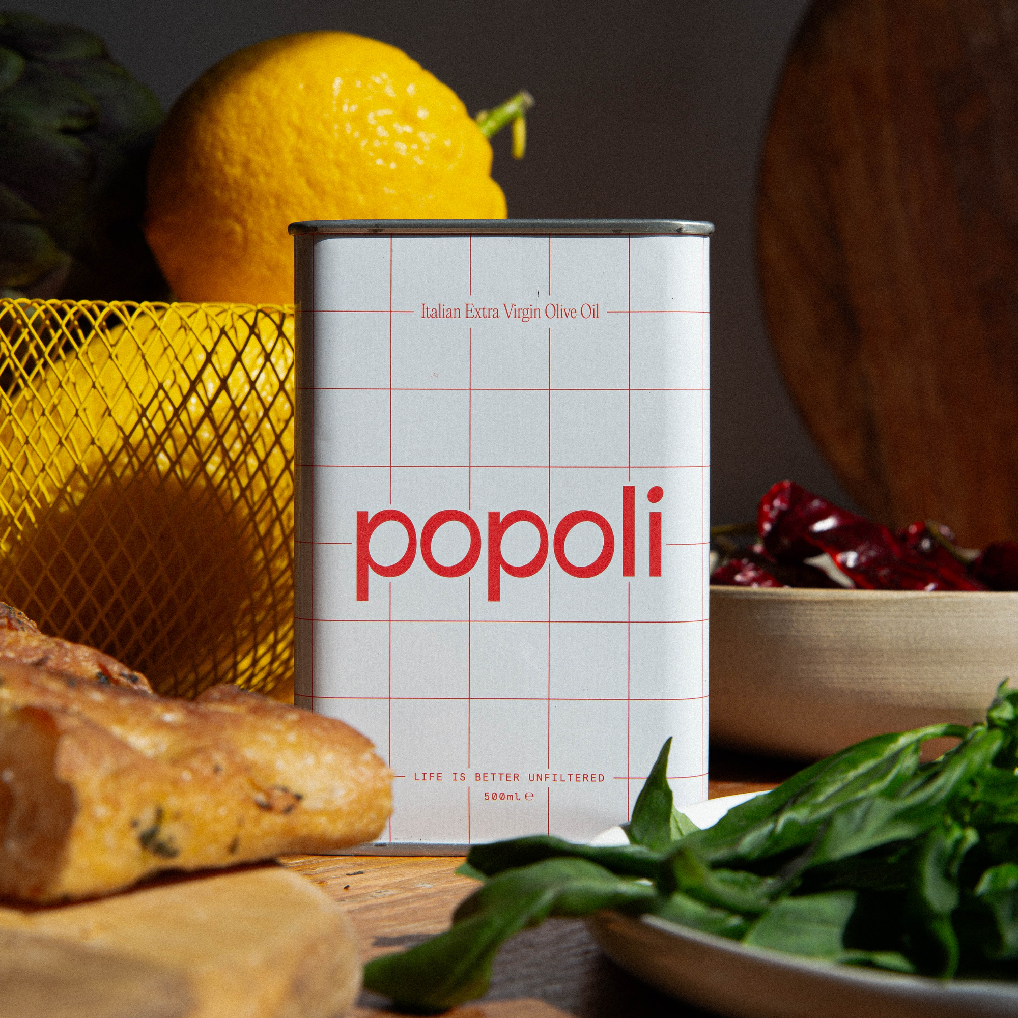 A can of Popoli Italian Extra Virgin Olive Oil stands on a wooden surface surrounded by fresh ingredients, including basil, bread, a lemon, and red chilies.