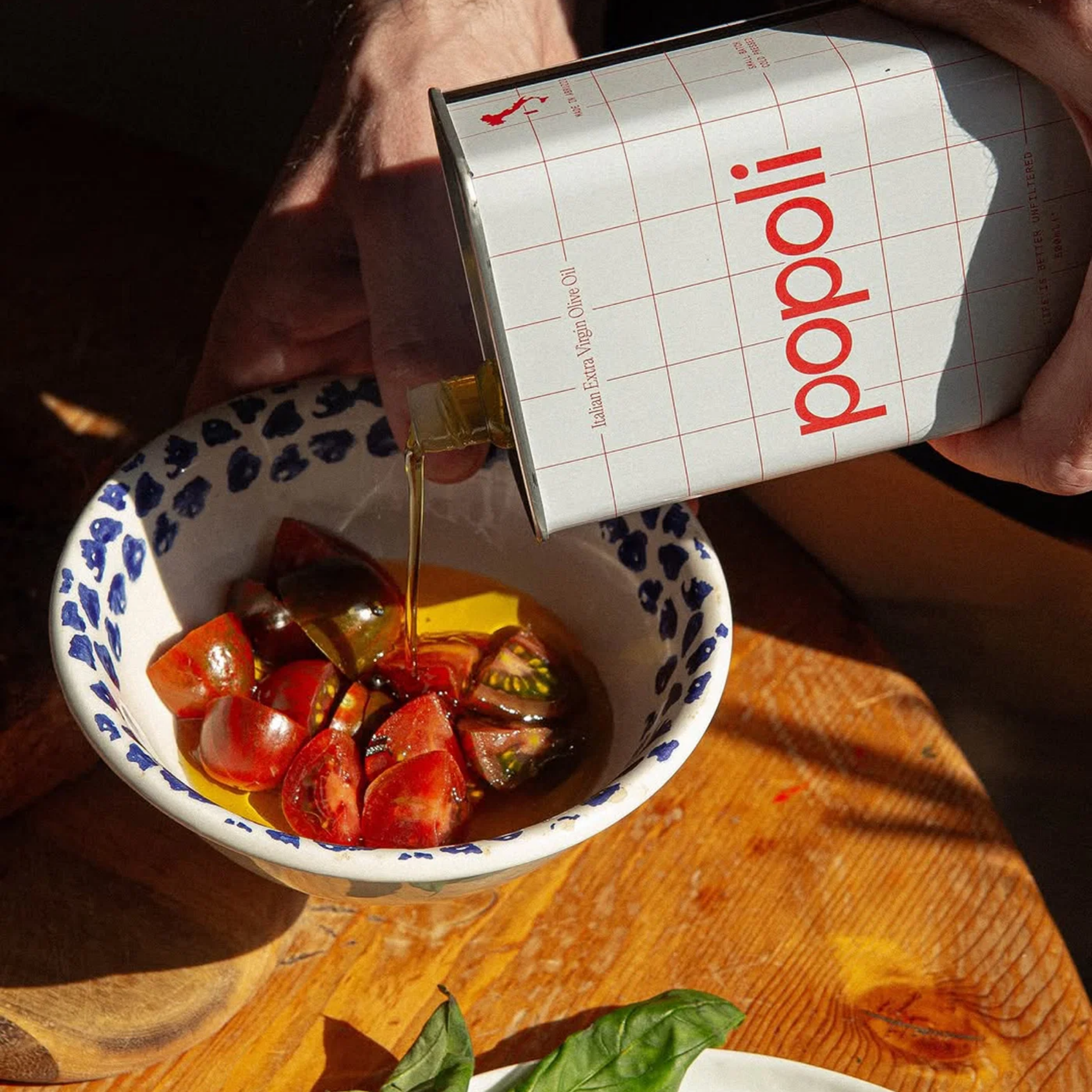 A hand holds a white can of Popoli olive oil over a bowl of sliced tomatoes and oil on a wooden table.