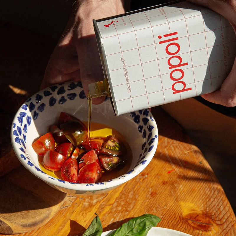 A hand holds a white can of Popoli olive oil over a bowl of sliced tomatoes and oil on a wooden table.