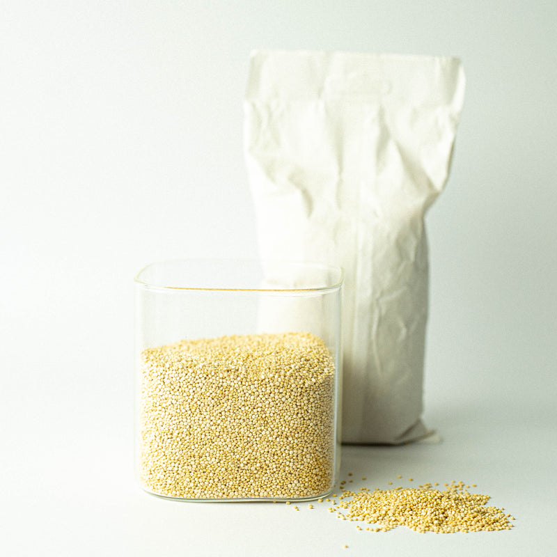 A glass jar filled with organic white quinoa in front of a recyclable paper bag, with a small pile of quinoa grains scattered in the foreground.