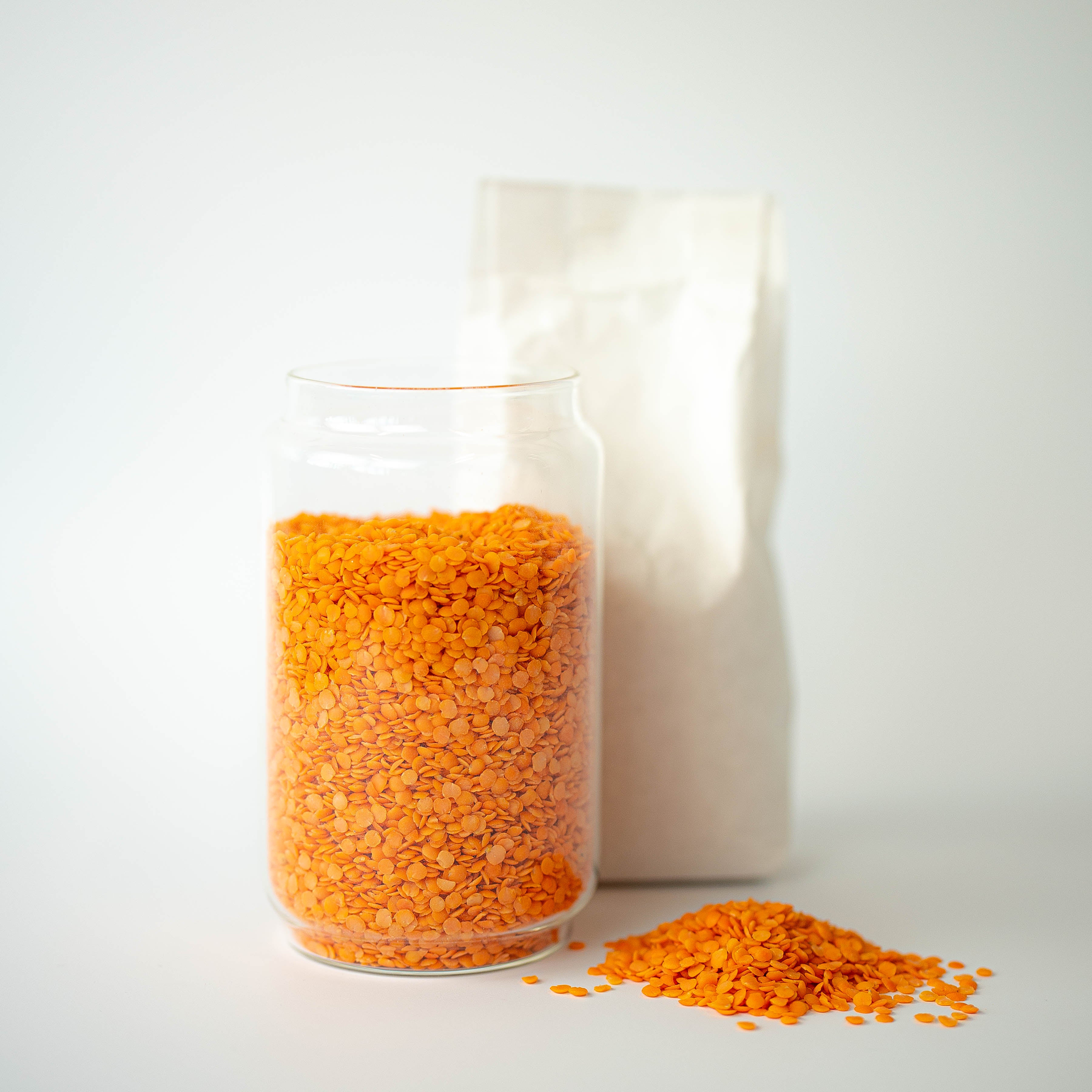 A clear glass jar filled with organic red split lentils, with a recyclable paper bag in the background and a small pile of lentils on the foreground.