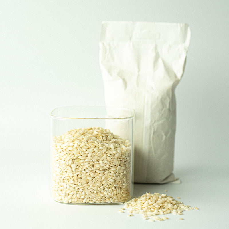 A glass jar filled with Arborio rice in front of a recyclable paper bag with rice, against a white background.