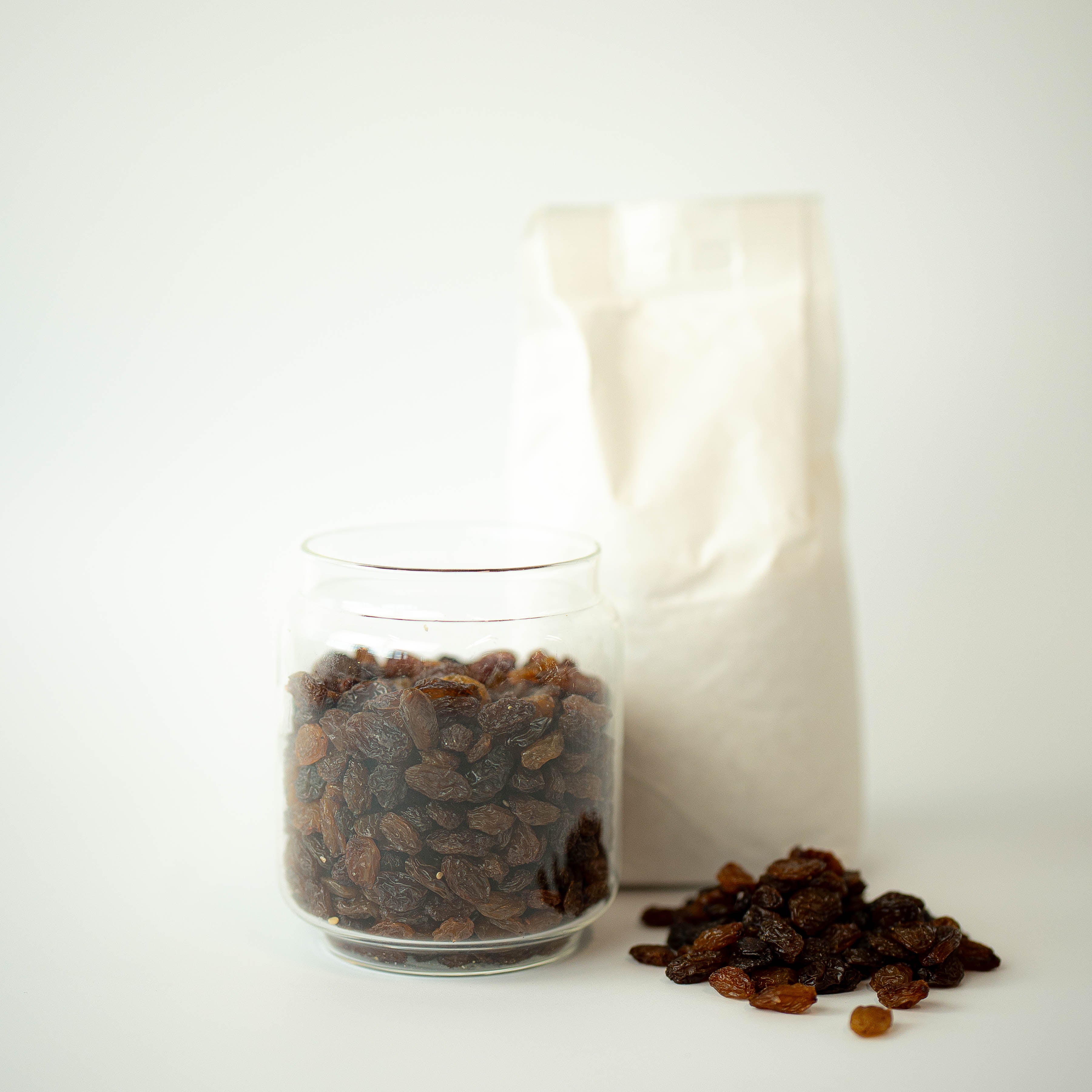 A glass jar filled with sultanas (dried raisins) in front of a recyclable paper bag, with a small pile of sultanas scattered in front.