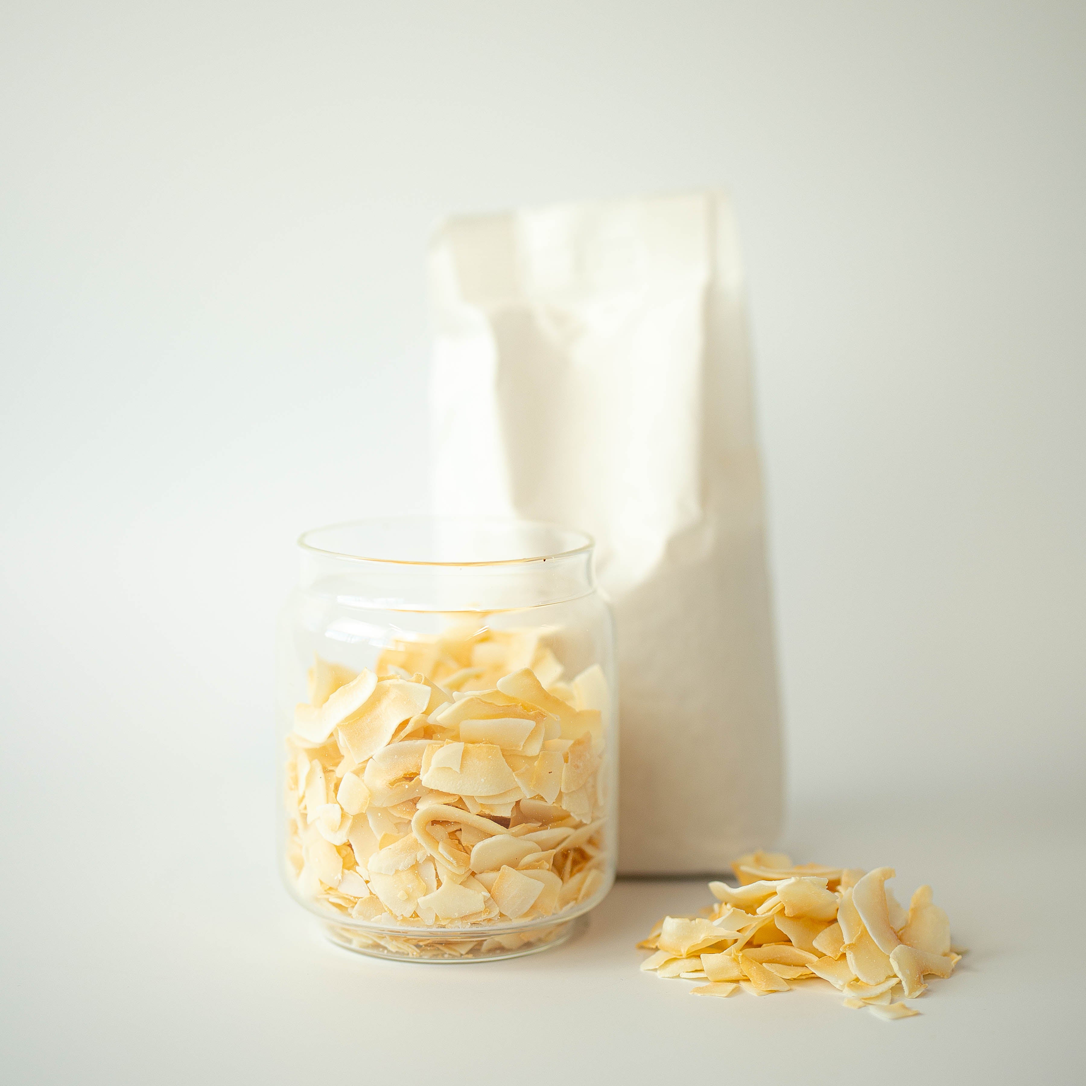 A glass jar filled with toasted coconut chips in front of a recyclable paper bag, with some chips scattered in the foreground.